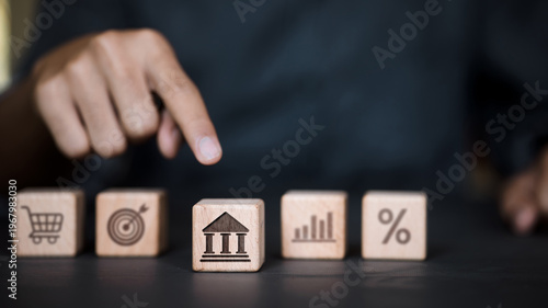 Hand pointing at bank icon on wooden block among financial and business symbols.