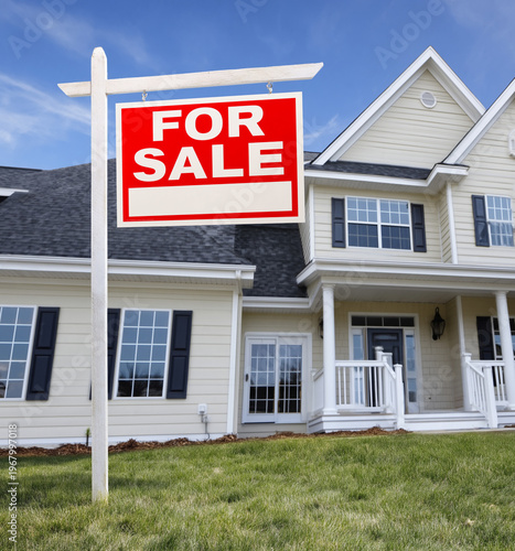 A large two story modern suburban home with cream siding dark shutters and white trim stands on a lush green lawn under a clear blue sunny sky with a 