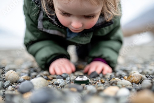 A young child in a green jacket closely examines a small object on a pebble-covered ground.
