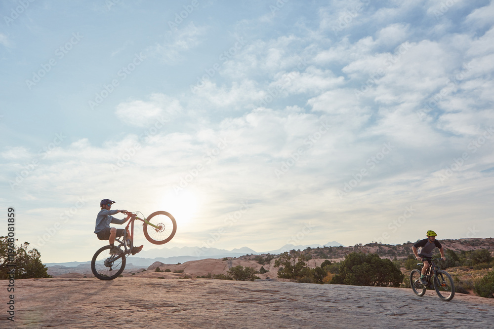 Fototapeta premium Man, mountain bike and wheelie in summer with friends, sky and extreme sports for trick with balance. People, bicycle and sunshine with ride, exercise and outdoor on hills with space in countryside