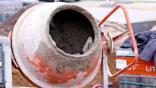Worker spraying water into a concrete mixer drum, mixing cement and aggregate, with visible construction site background 