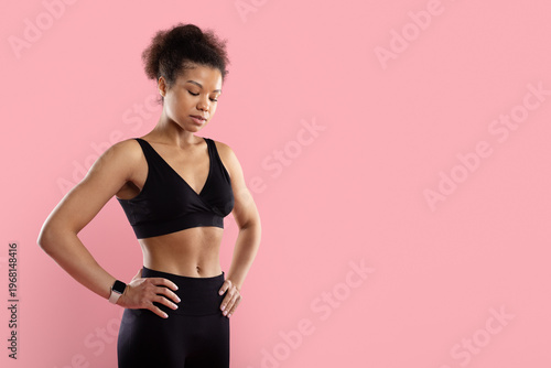 Focused Black sportswoman in black sports bra standing with hands on hips looking down on pink studio background. Thoughtful fitness portrait with copy space