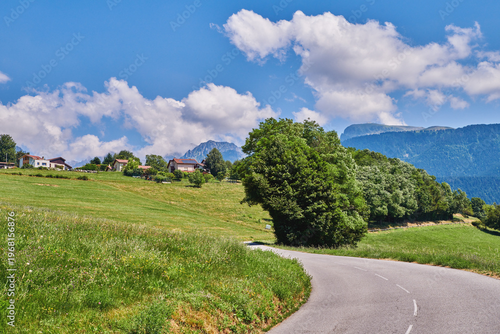 Obraz premium Road, countryside and farmland in nature with field, sky and clouds with mountains, trees and horizon. Outdoor, grass and buildings on hill with street, infrastructure or asphalt in springtime