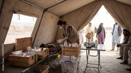 A female medical worker wearing a mask and gloves provides care to a patient lying on a cot inside a temporary medical tent, with other people waiting in the background.