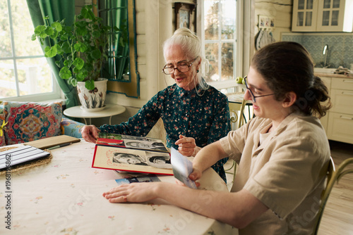 Senior Caucasian woman showing photo album to young adult Caucasian grandson at table, both sitting and looking at old photographs, engaging in sharing family memories together