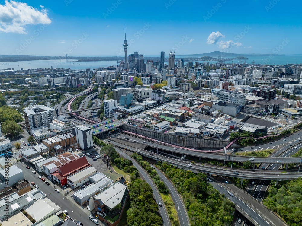 Obraz premium Aerial view of Auckland, New Zealand, showcasing the city skyline with the Sky Tower, buildings, and harbor. Cars travel on the highway, illustrating urban transportation.