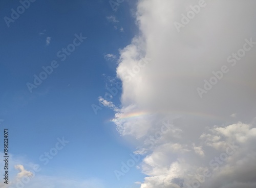 Cumulus cloud, blue sky and rainbow. The sky is divided into a cloudy part and a clear blue sky. A rainbow is partially visible in the center of the frame. Weather forecast, partly cloudy.