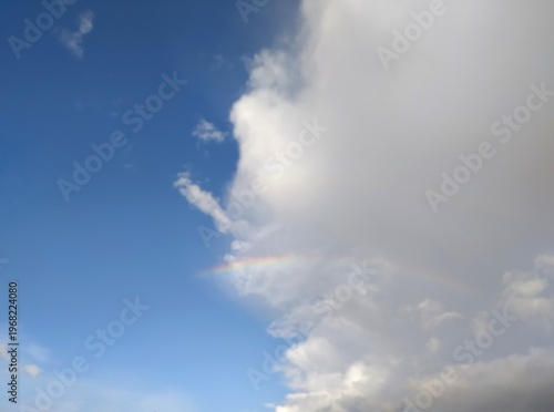 The sky is divided into a cloudy part and a clear blue sky. A rainbow is partially visible in the center of the frame. Weather forecast: partly cloudy. Cumulus cloud against a blue sky.
