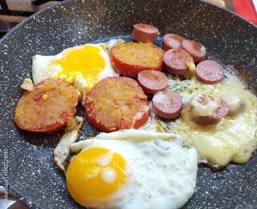  Fried eggs witр sausages, cheese and breaded tomatoes in a pan on the stove, close-up, isolated, breakfast, home cooking.