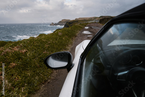 White car driving on rugged coastal cliff road with stormy Atlantic Ocean waves, Azores, Portugal