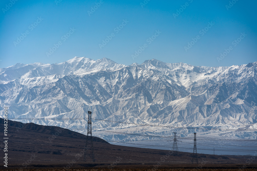 Obraz premium Snow covered Qilian Mountains in China with power lines in the foreground under a clear blue sky Snow covered mountains rise majestically behind a rugged landscape with power lines 