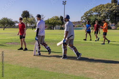 Male teammates in shorts and shirts walking across pitch at park carrying cricket bats and pads
