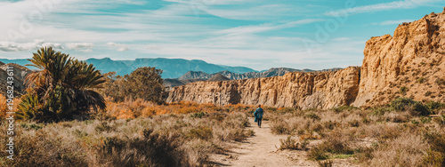 Fotografie Woman walks on path in mountainous semi desert landscape with vegetation and palm trees under a blue sky with some thin clouds