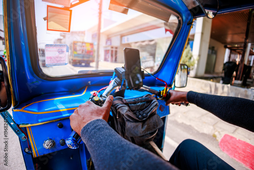 Point of view from behind a driver operating a blue Tuk Tuk taxi on a city street during a sunny day in Chiang Mai, Thailand.