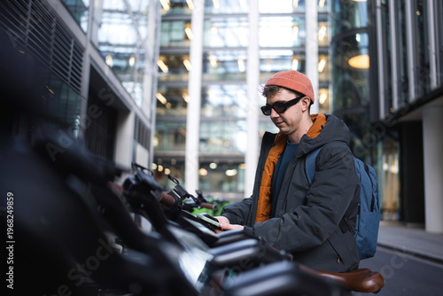 Canvas Print Young man wearing sunglasses and a beanie hat unlocking an electric scooter from