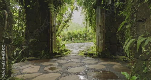 Ancient stone gate draped in moss and ferns opens pebbled path, inviting to serene, secret tropical garden in dense jungle foliage. Travel to Indonesia, explore Bali culture. Natural background
