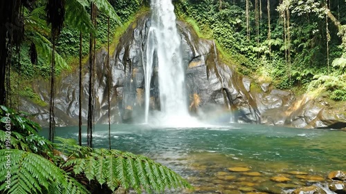 A majestic waterfall cascades down a rocky cliff face into a clear turquoise pool surrounded by lush green tropical vegetation and hanging vines