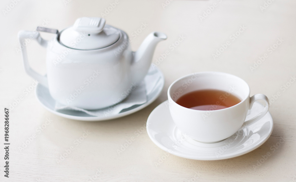 Fototapeta premium A white teapot and a cup of hot tea on a light surface