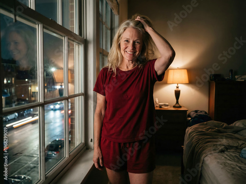 Smiling mature woman in red pajamas standing by the window at night, cozy home atmosphere