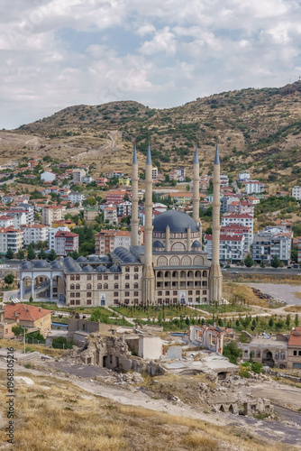 Turkey. Vertical scene showing a large mosque with minarets and a smaller dome, set against a backdrop of residential buildings and hills in Turkey