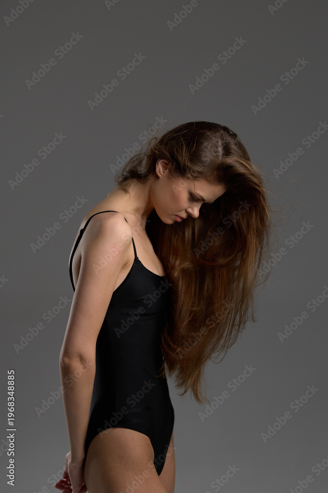 Fototapeta premium Woman with long hair in black bodysuit looking down, posing against gray background with soft lighting.