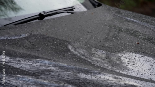 Close-up of raindrops on dark car hood during rainy weather. Water beads and streaks create reflective textured surface with windshield wiper in background. Moody outdoor scene, shallow depth of field