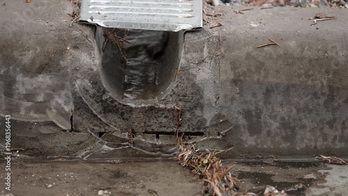 Rainwater flowing from a metal street drain into a concrete gutter. Small debris and pine needles gather at the outlet as water trickles across a wet pavement. Urban drainage detail after rainfall.