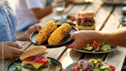 Hands passing grilled corn at outdoor barbecue table with burgers and salad in warm evening light