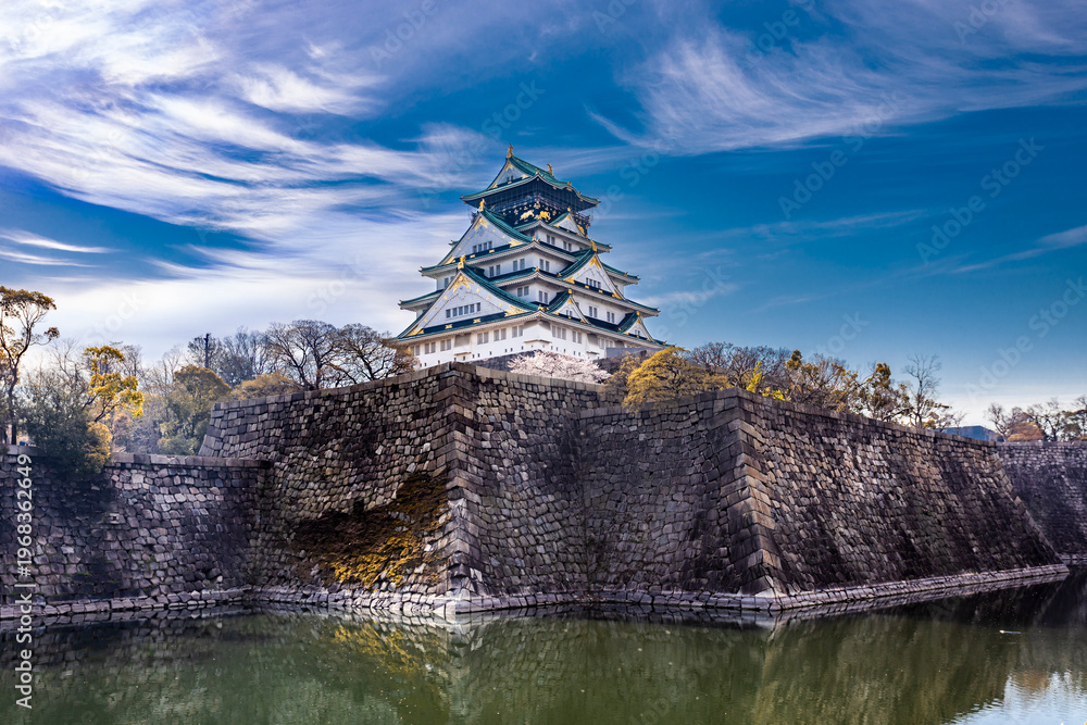 Fototapeta premium Magnificent view of Osaka Castle in spring, Japan. The historic fortress is surrounded by stone walls, a peaceful moat, and blooming cherry blossoms under a vibrant blue sky in early April.