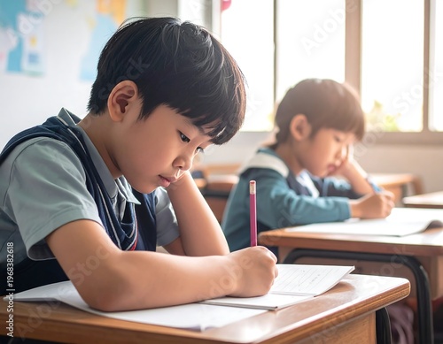 Two young students, both Asian, focus intently on their written assignments in a well-lit classroom setting. One rests his head