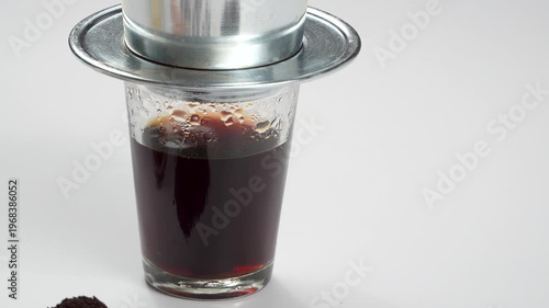 Wide rotating view of a full glass of hot Vietnamese black coffee with a metal drip filter on top against a white background. Commercial cafe footage.