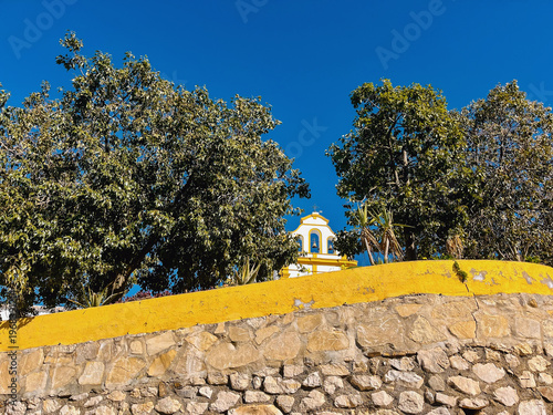 Old church tower between trees behind a stone wall under a clear blue sky. Colmenar, Malaga, Andalusia, Spain.