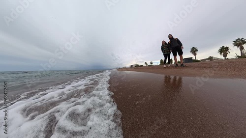 A slow-motion shot with a rotating camera captures a man and a woman standing on a sandy beach. The waves gently approach the shore, creating a serene and dynamic scene with a wide-angle view of the c