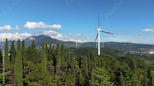 Aerial wide shot of a white wind turbine spinning against a backdrop of the Italian Alps and a clear blue sky.