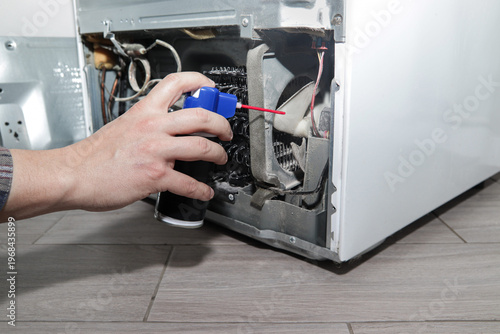 Male hand spraying lubricant on refrigerator condenser and internal fan. Concept of fridge maintenance, refrigerator servicing, appliance care, lubrication, technical maintenance.