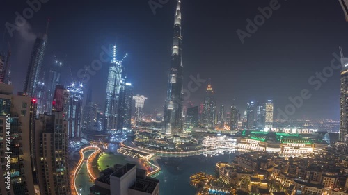 Tall skyscrapers rising above the famous mall and fountain in Dubai Downtown. Aerial panoramic view during all night time-lapse showing city lights slowly turning off.