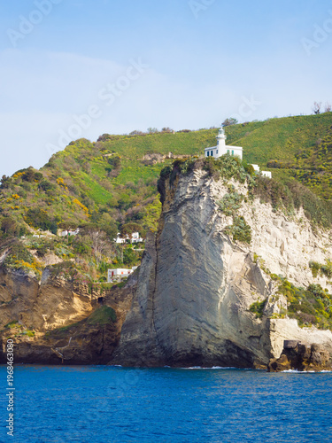 White lighthouse stands on top of vertical rock cliff above Mediterranean sea. Green hills and houses are in background under clear blue sky. Scenic coastal landscape of Phlegraean Fields region