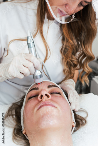 Slika na platnu A professional aesthetician in a white uniform uses a radiofrequency skin tightening device on a woman's forehead during a rejuvenating facial procedure in a wellness spa clinic