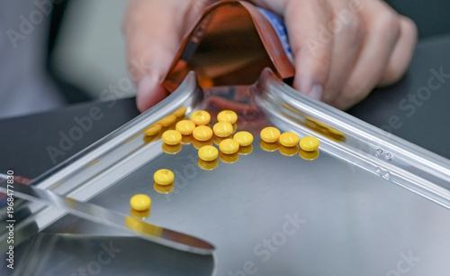 Fotografie Yellow tablet pills on stainless steel tray with blurred pharmacist hand counting medication into plastic zipper bag in pharmacy showing prescription dispensing accuracy safety and healthcare process