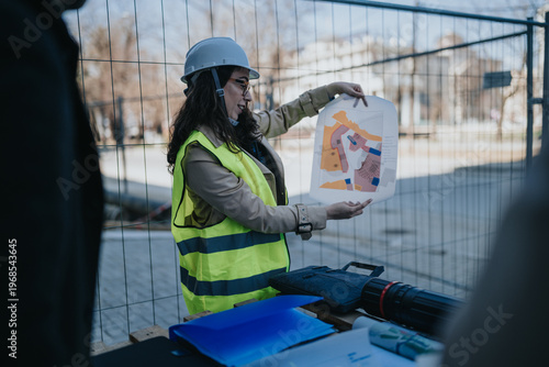 A construction engineer in a high-visibility vest and hard hat holds and examines a site plan. The scene shows an engineer presenting building plans at a fenced construction site.