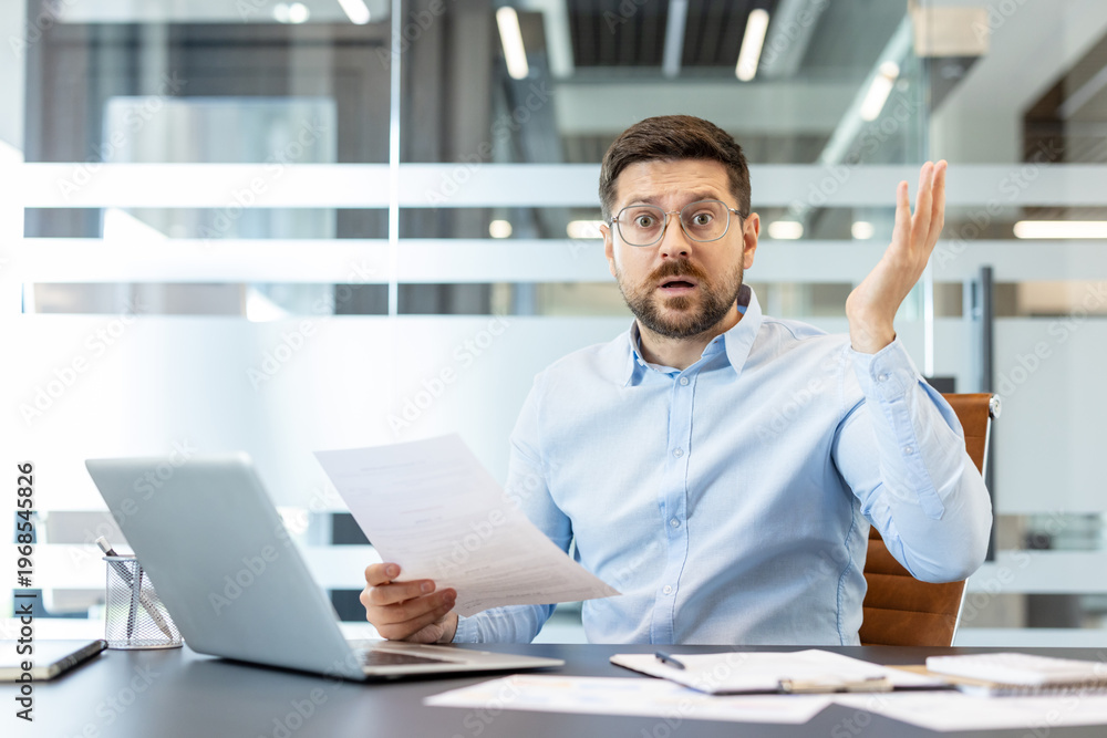 Obraz premium Stressed male employee holding a document, looking confused and frustrated with an open mouth gesture, sitting at his office desk with a laptop and papers
