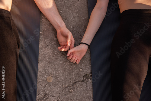Couple lying on yoga mats holding hands on concrete, top-view moment of connection and shared wellness during outdoor fitness, calm, supportive partnership and relaxation