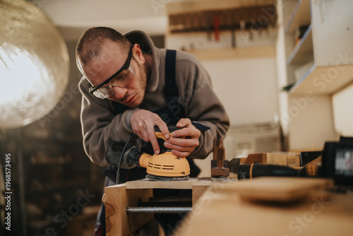 Foto A male carpenter sands a wooden plank with an electric sander while wearing safety goggles