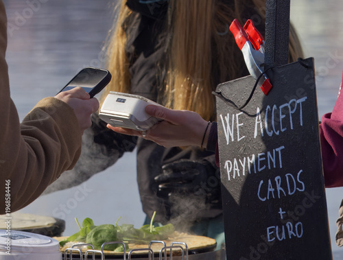 Perfect for modern finance and business themes. A customer uses a smartphone for contactless payment at a popular Naplavka market street food stall with a sign accepting cards and Euro.