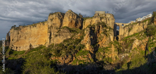 View of Ronda with its cliffs and buildings under a cloudy sky near the famous bridge at sunset