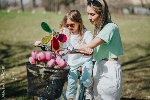 A mother helps her young daughter ride a bicycle while holding the handlebars. The pair ride together in a sunny park with tulips in the bike basket.