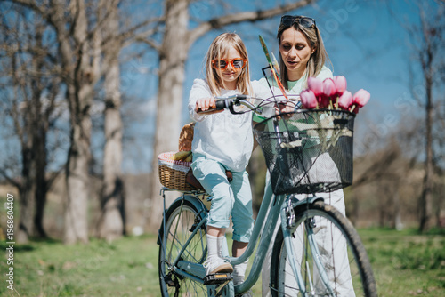 A mother helps her young daughter ride a bicycle while standing beside her in a park. They enjoy a sunny spring day with tulips in the bike basket.