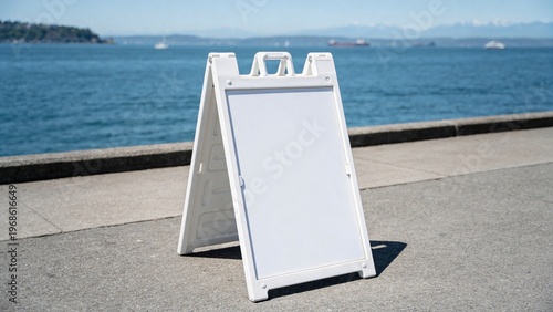 A blank white A-frame sign stands on a concrete walkway by the waterfront. The ocean and distant mountains are visible in the background under a clear blue sky.