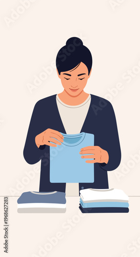 A woman with a dark bun hairstyle stands folding a light blue t-shirt on a table