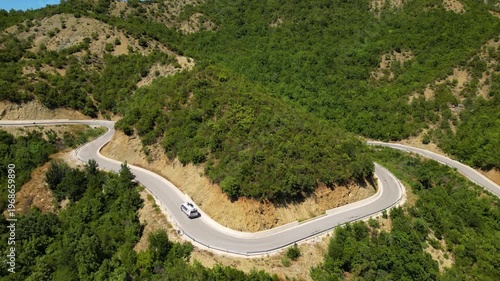 Aerial view of a campervan traveling, driving the iconic SH75 mountain road in Albania, winding through the rugged landscape above the vibrant turquoise waters of the Vjosa River in the summer sun.